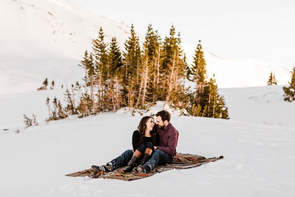 couple sitting on a Pendleton blanket during their snowy Colorado winter engagement photos in the Rocky Mountains