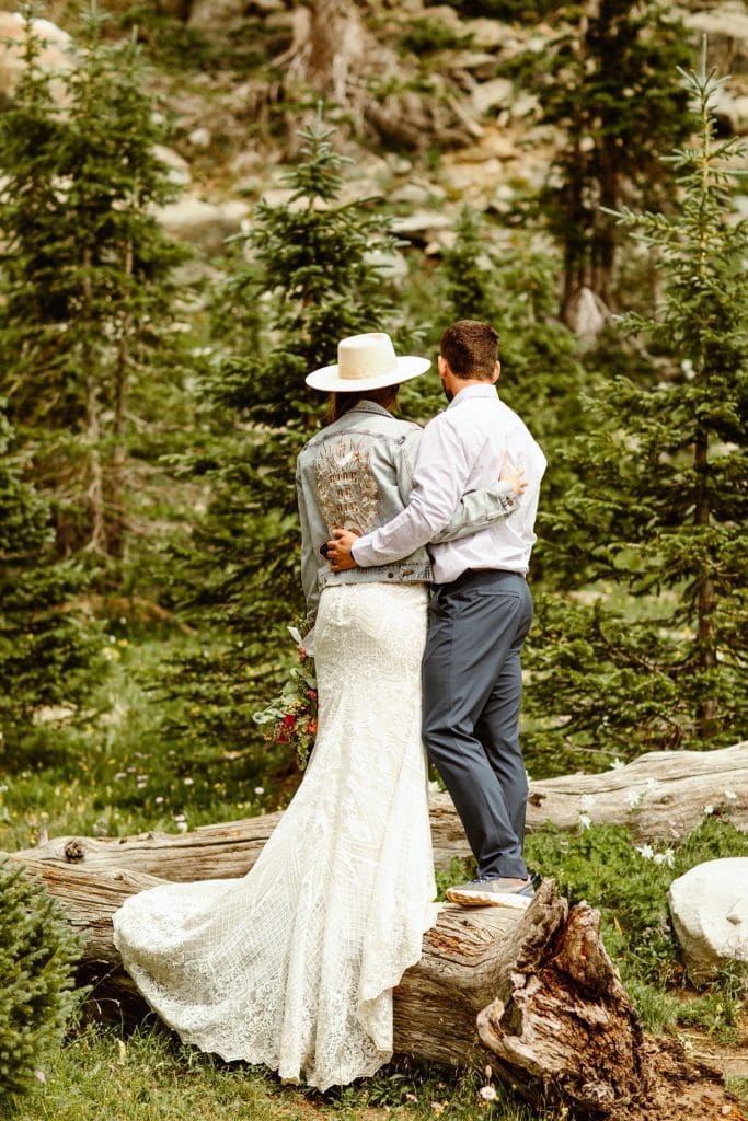 bride and groom standing together on a log in the woods of the Snowy Range Mountains after their Wyoming elopement - bride is wearing Rue de Seine denim jacket