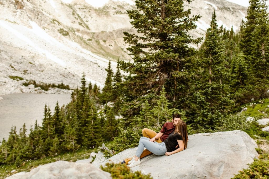 couple sunbathing on a rock together by the lake while having their mountain engagement photos taken