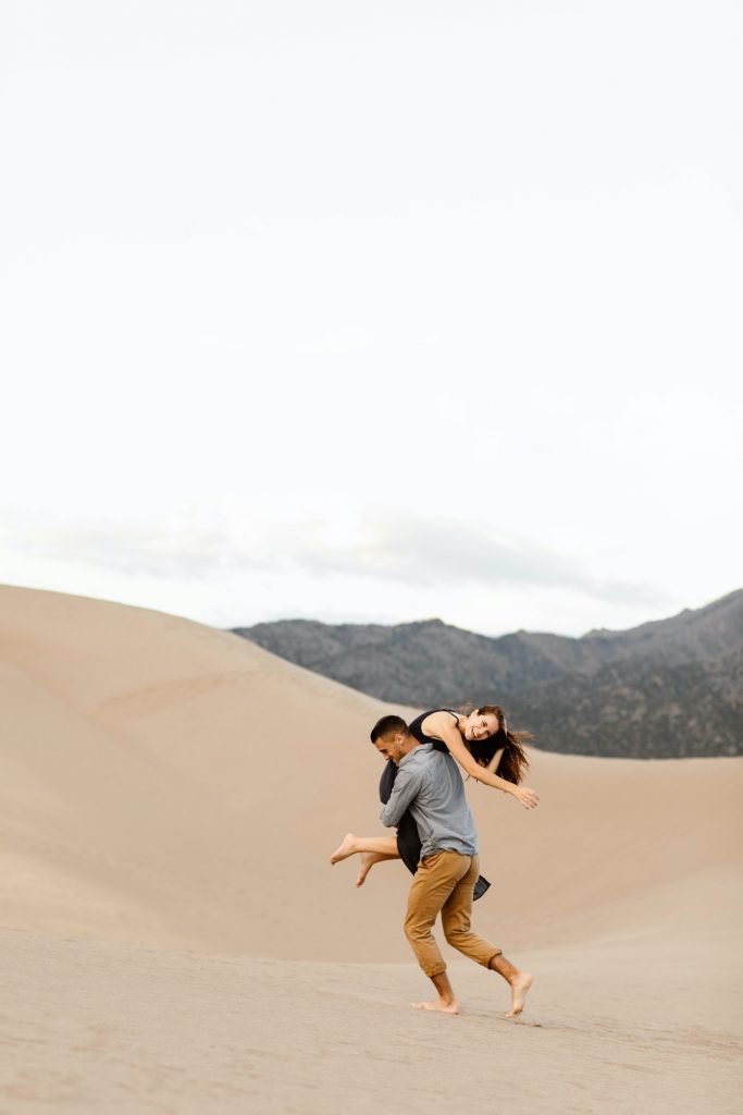 guy throwing girl over his shoulder at the sand dunes with mountains in the background
