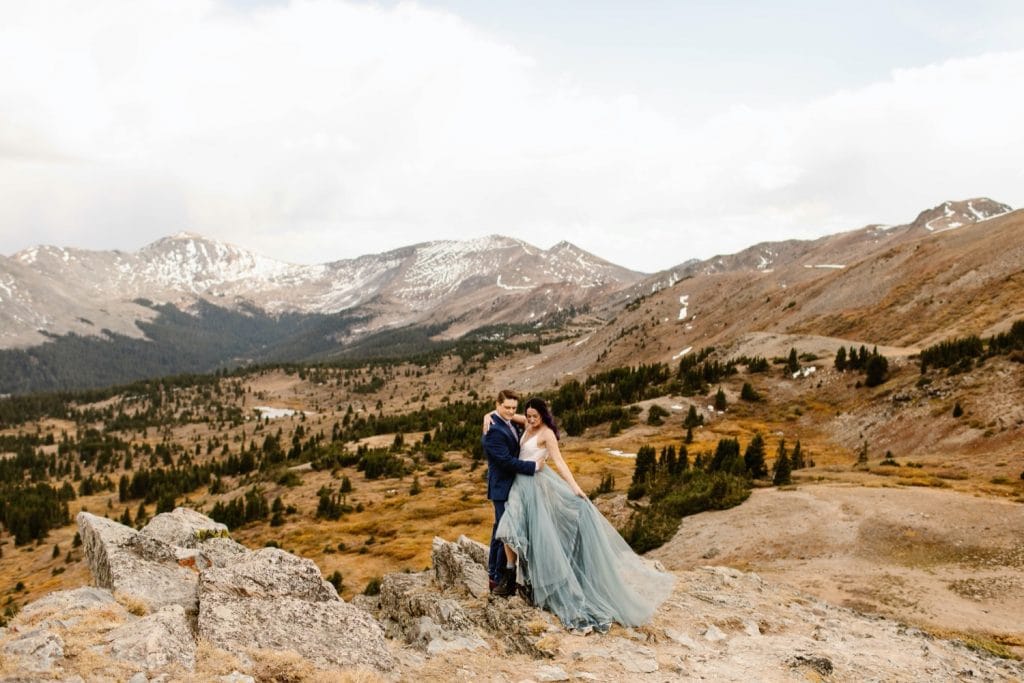 couple standing on the edge of a cliff in the mountains before their Buena Vista elopement ceremony | bride waving her dress in the wind