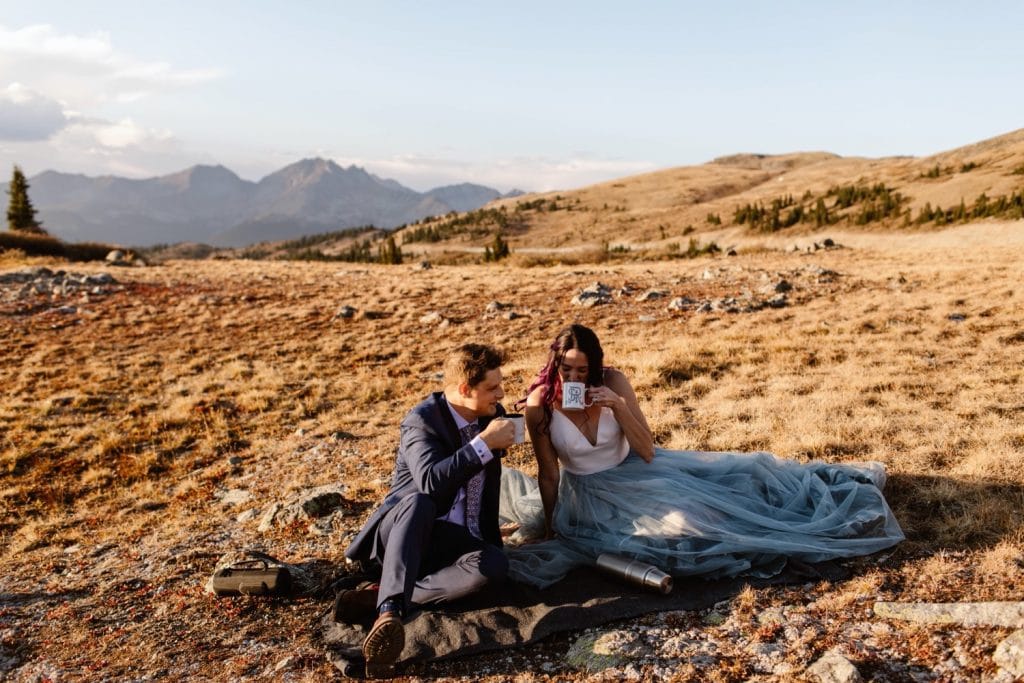 eloping couple drinking hot cocoa while sitting on a picnic blanket in the mountains