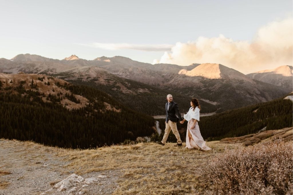 bride and groom walking hand in hand in the mountains while the smoke of a wildfire emerges from behind them