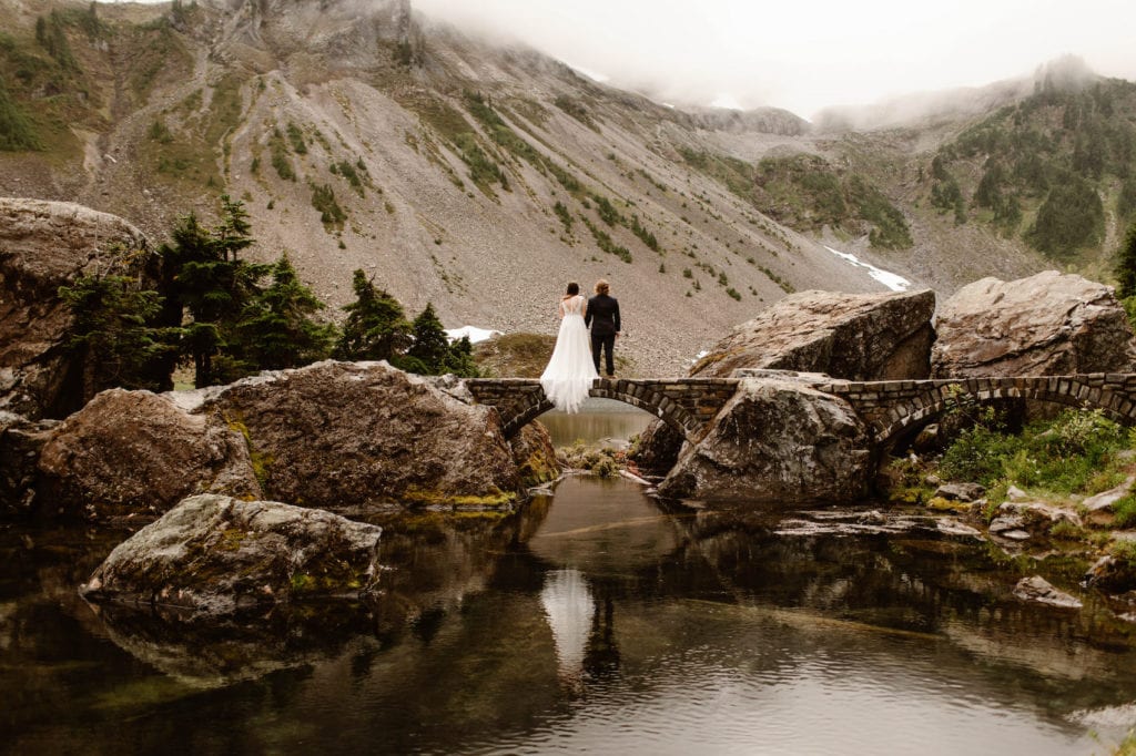 eloping couple standing on a lakeside bridge while the bridge wears a chiffon summer elopement dress