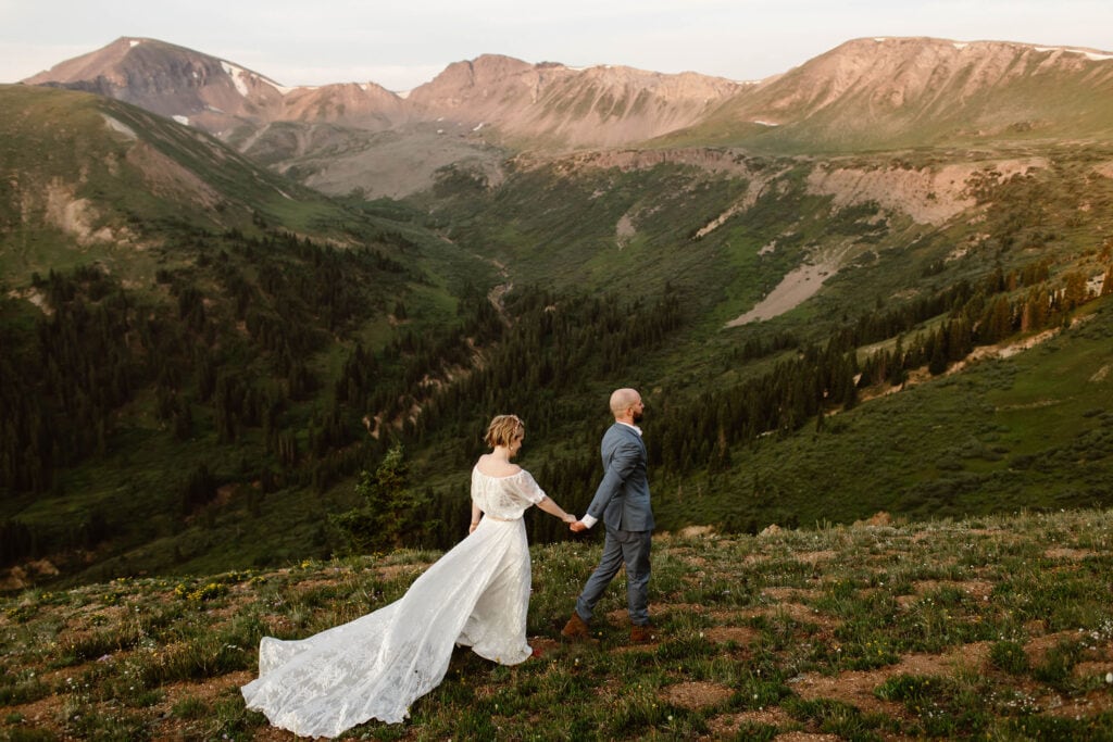 bride and groom walking in the mountains while the bride wears a wedding dress with a long lace train that she has chosen not to bustle