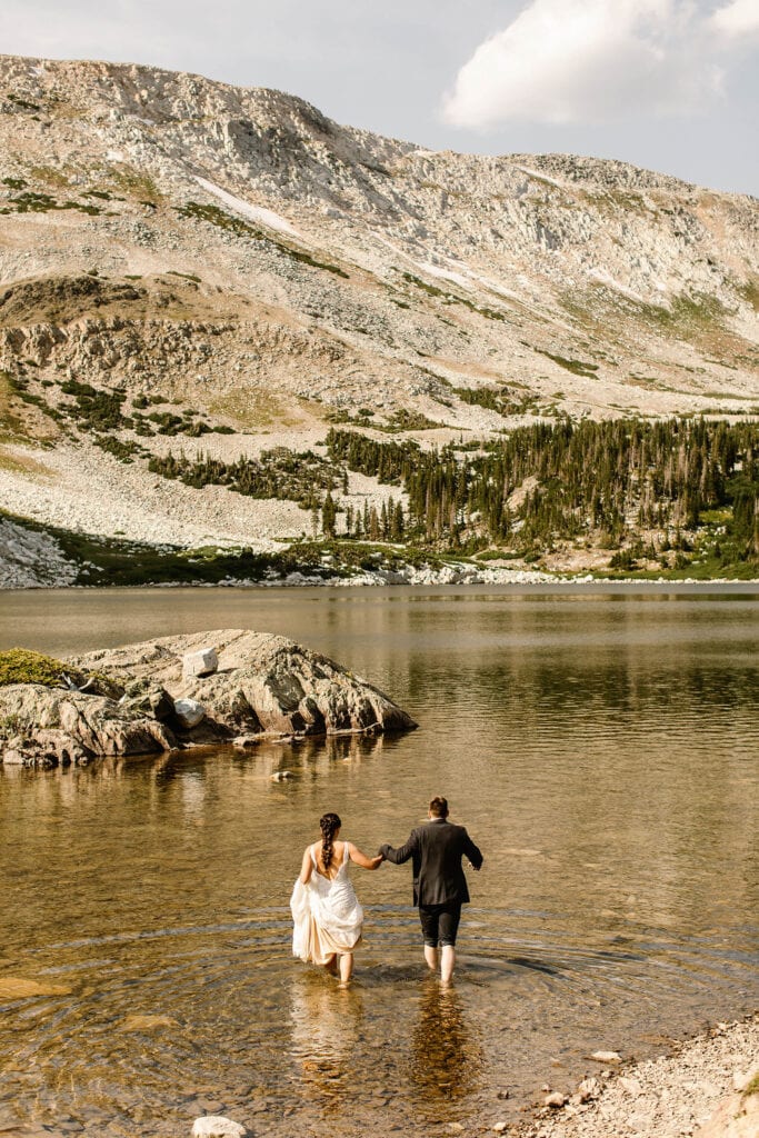 bride looping the train of her wedding dress around her wrist while she wades into a lake