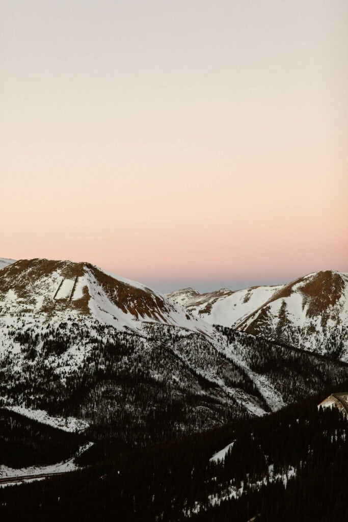 a layered pink and orange sunset over the snow dotted mountains from the overlook on Loveland Pass Colorado