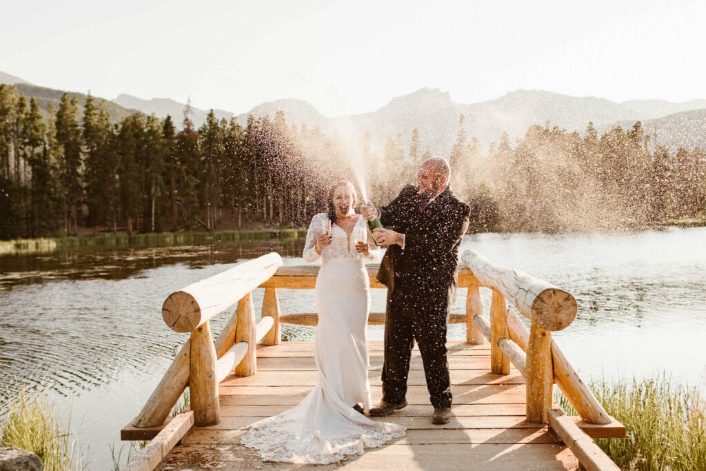 married couple popping champagne on the dock at Sprague Lake Trail in Colorado at sunset on their wedding day in the summer