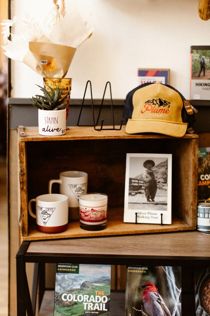 Colorado merchandise shelf at Plume Coffee Bar with a branded coffee shop hat and a Silver Plume themed black and white postcard alongside mugs and hiking guide books